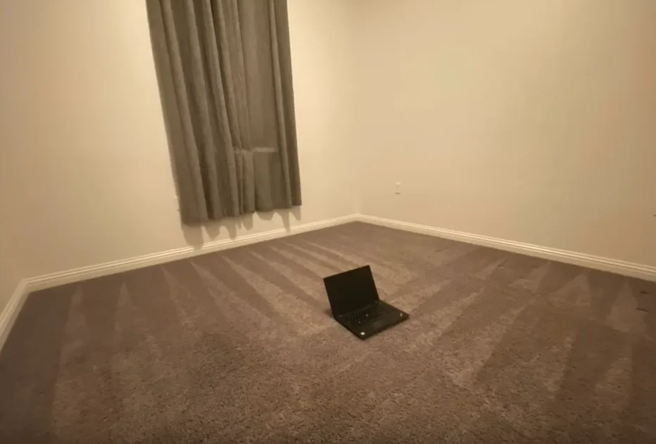 A black laptop sits open on a beige carpeted floor in an otherwise empty room with white walls and a single grey curtain…