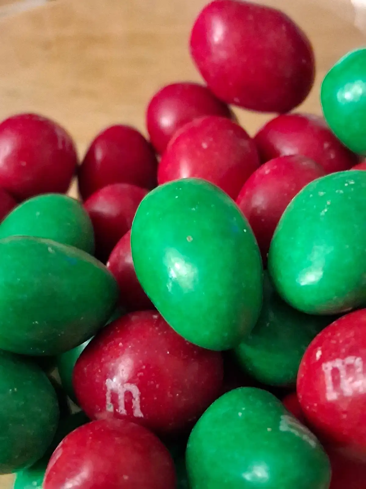 A close-up photo shows a pile of red and green M&M candies on a wooden surface, with some red ones showing the white 'm'…