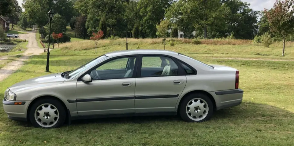 A silver Volvo sedan is parked on a grassy lawn with trees and a dirt road in the background. The overlaid text reads 'i…