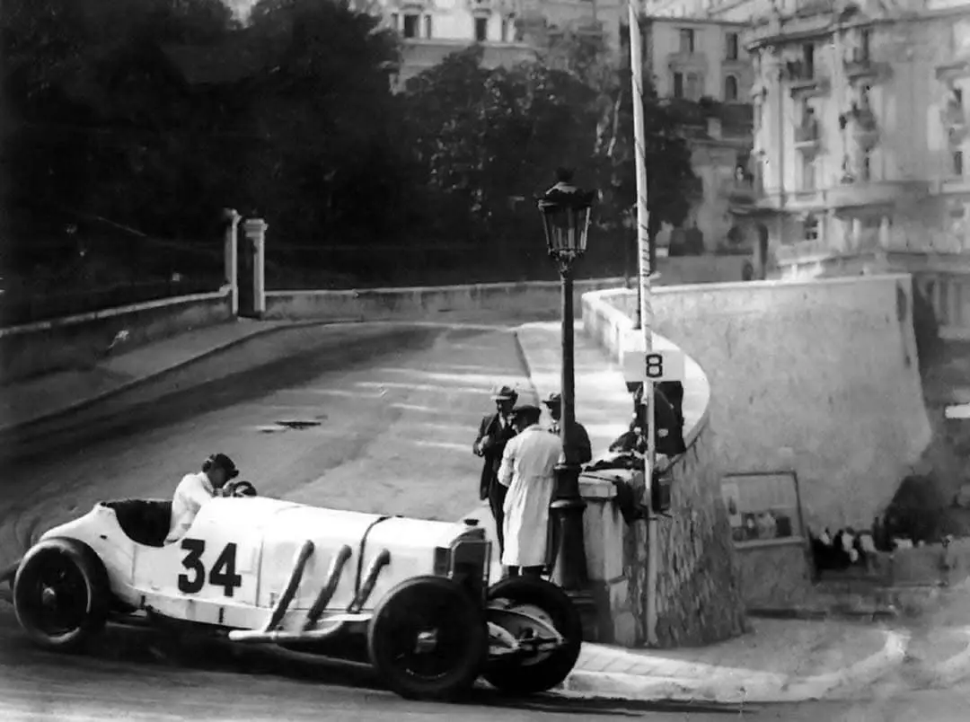 A vintage black-and-white photo shows a 1920s open-wheel race car, numbered 34, navigating a curved road with spectators and…