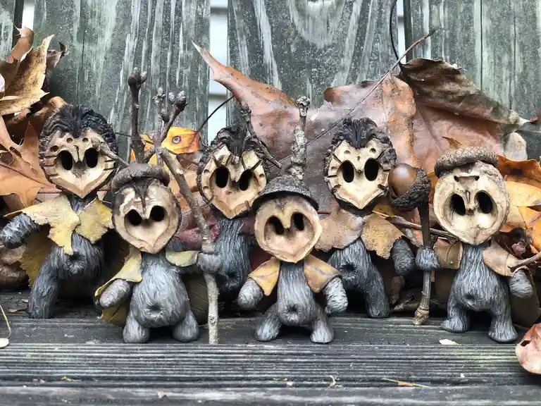 Five small figurines made from acorn caps and twigs, dressed in leaf cloaks and holding sticks, stand in a row on a wooden…