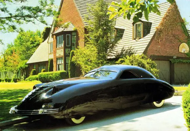 a glossy black 1939 darrin sportster, a rare and iconic american automobile, is parked on a driveway in front of a large…
