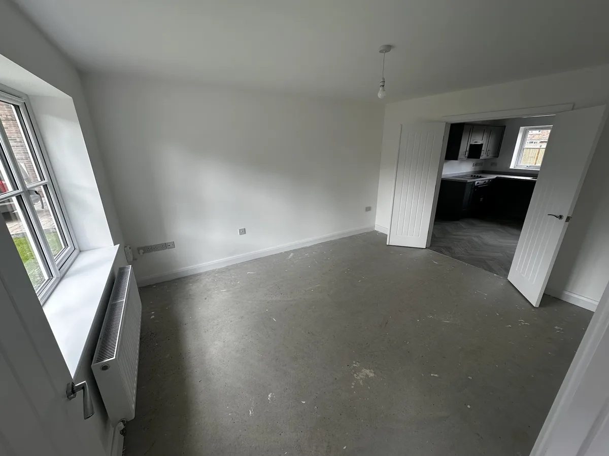 An empty, unfurnished room with bare concrete floors and white walls, featuring a window with a radiator below and a doorway…