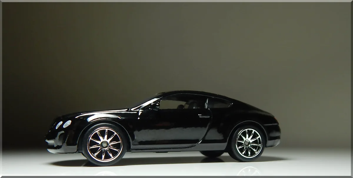 A glossy black toy model of a luxury sports car, likely a Bentley, is shown in profile against a plain gray background. The…