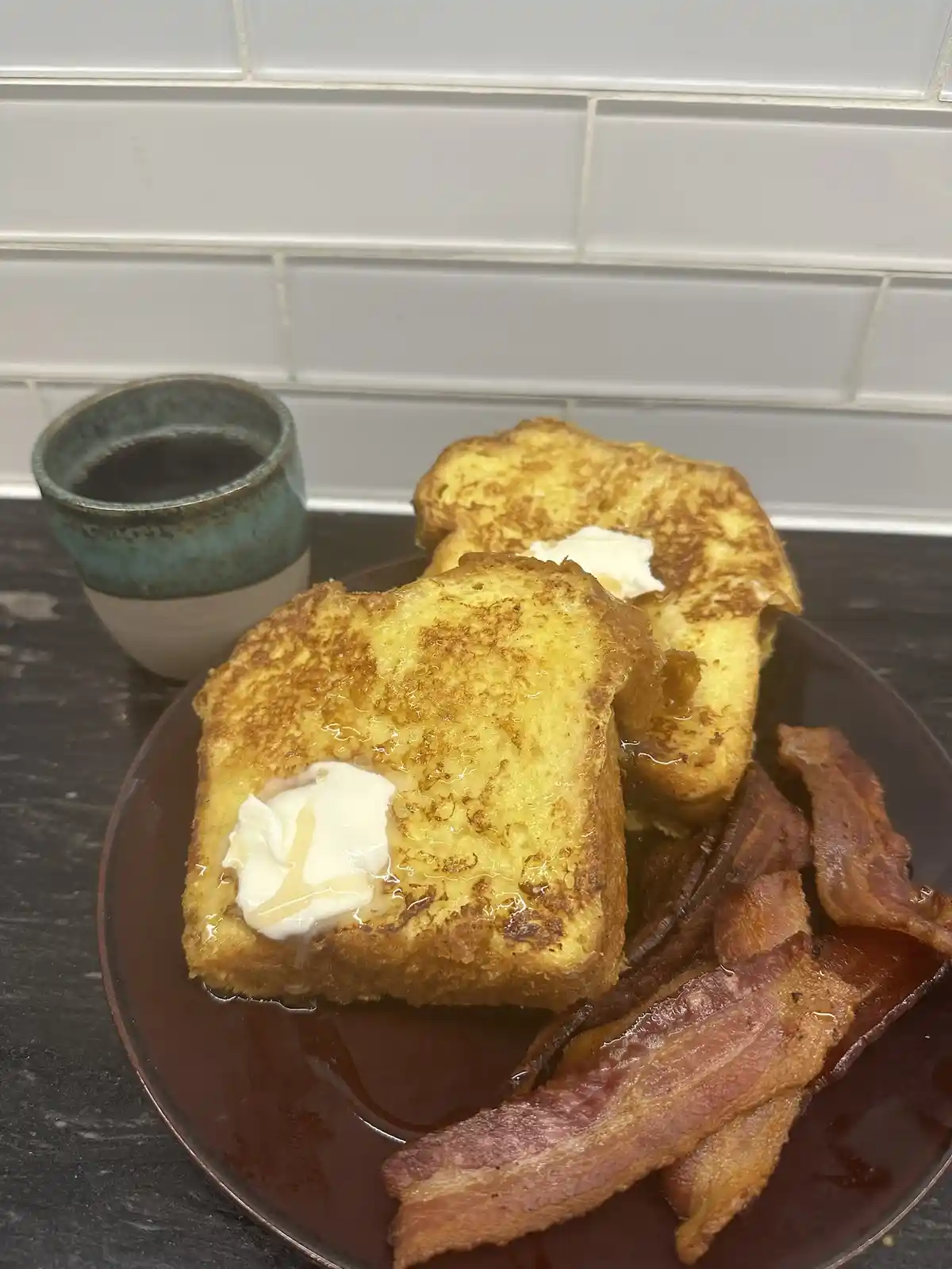 A plate of golden-brown French toast with butter and syrup, accompanied by crispy bacon and a cup of coffee, set against a…