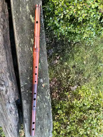 A wooden flute with a rich, swirling grain pattern rests vertically on a weathered, mossy wooden plank outdoors, next to…