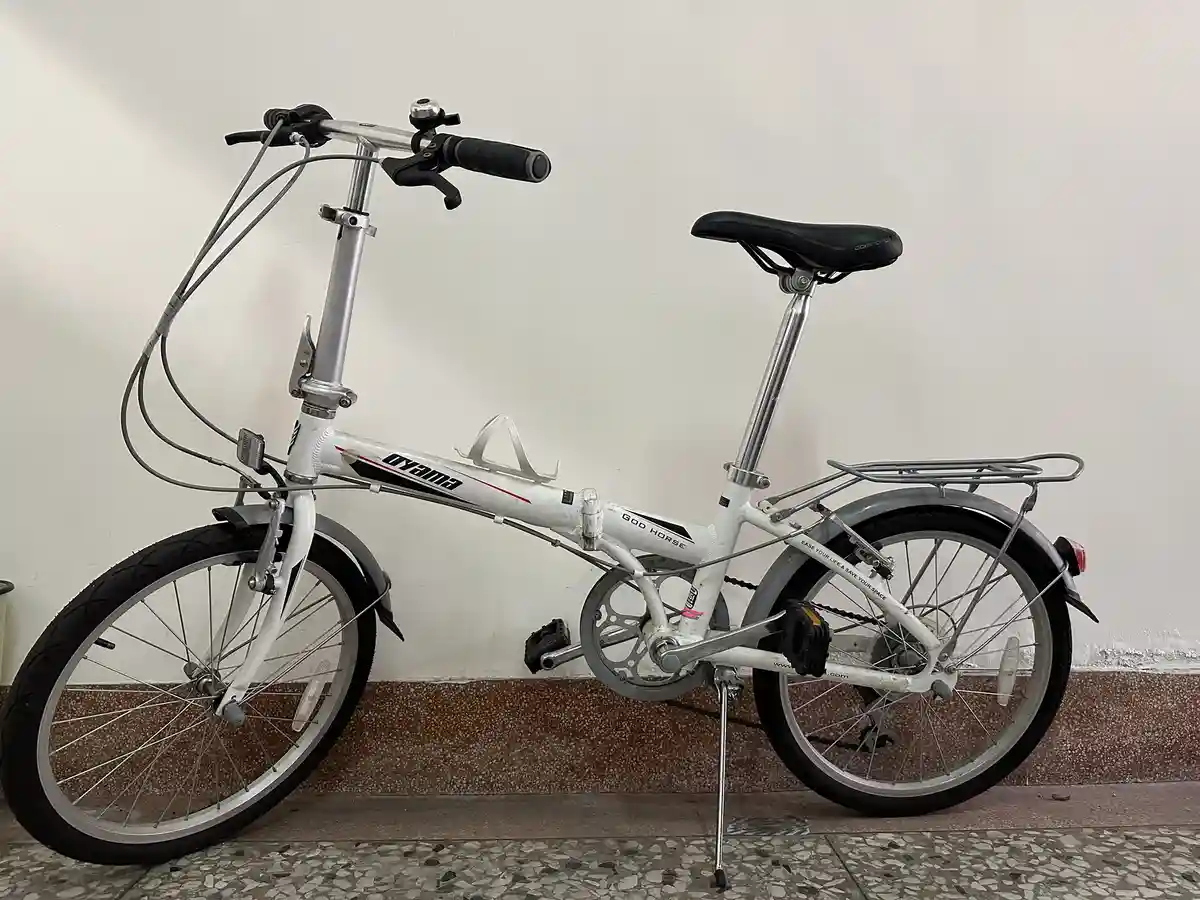 A white folding bicycle with 'god horse' written on the frame stands upright on a kickstand against a plain wall. The bike…