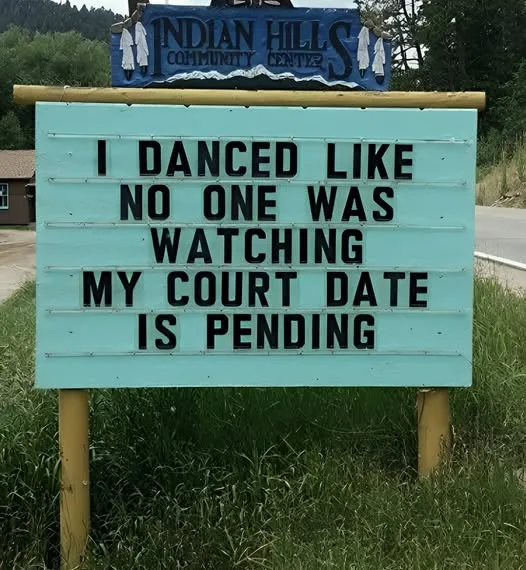A light blue outdoor sign with black lettering reads 'I DANCED LIKE NO ONE WAS WATCHING MY COURT DATE IS PENDING'. Above it…