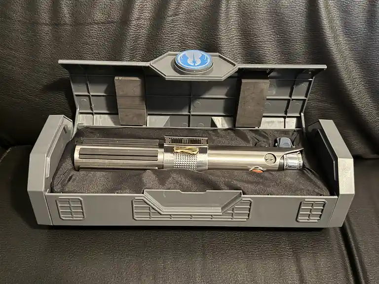 A silver toy lightsaber with gold accents rests horizontally in a grey plastic display case lined with black fabric, which…