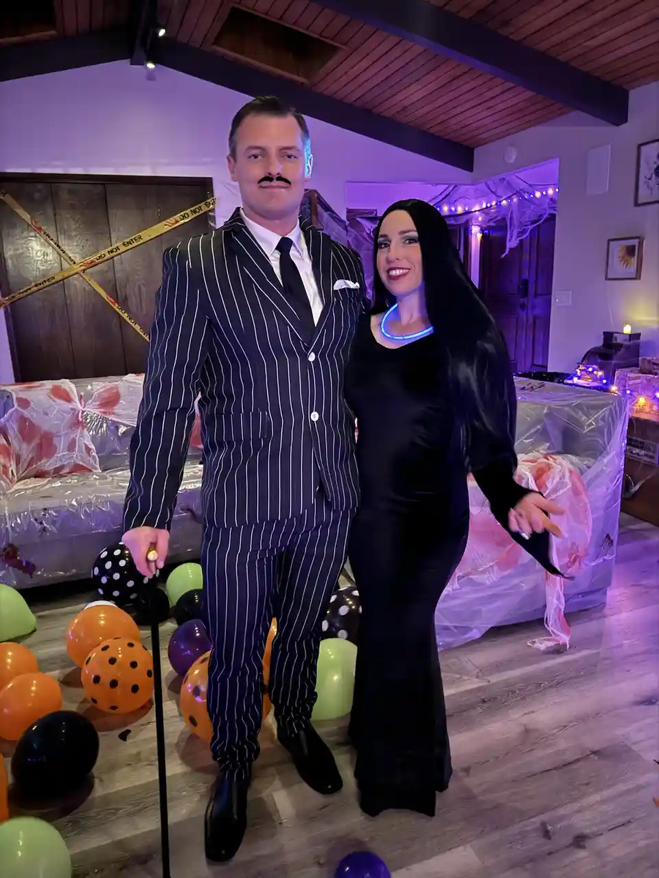 A man and woman dressed as the Addams Family characters Gomez and Morticia pose for a photo in a Halloween-decorated room…