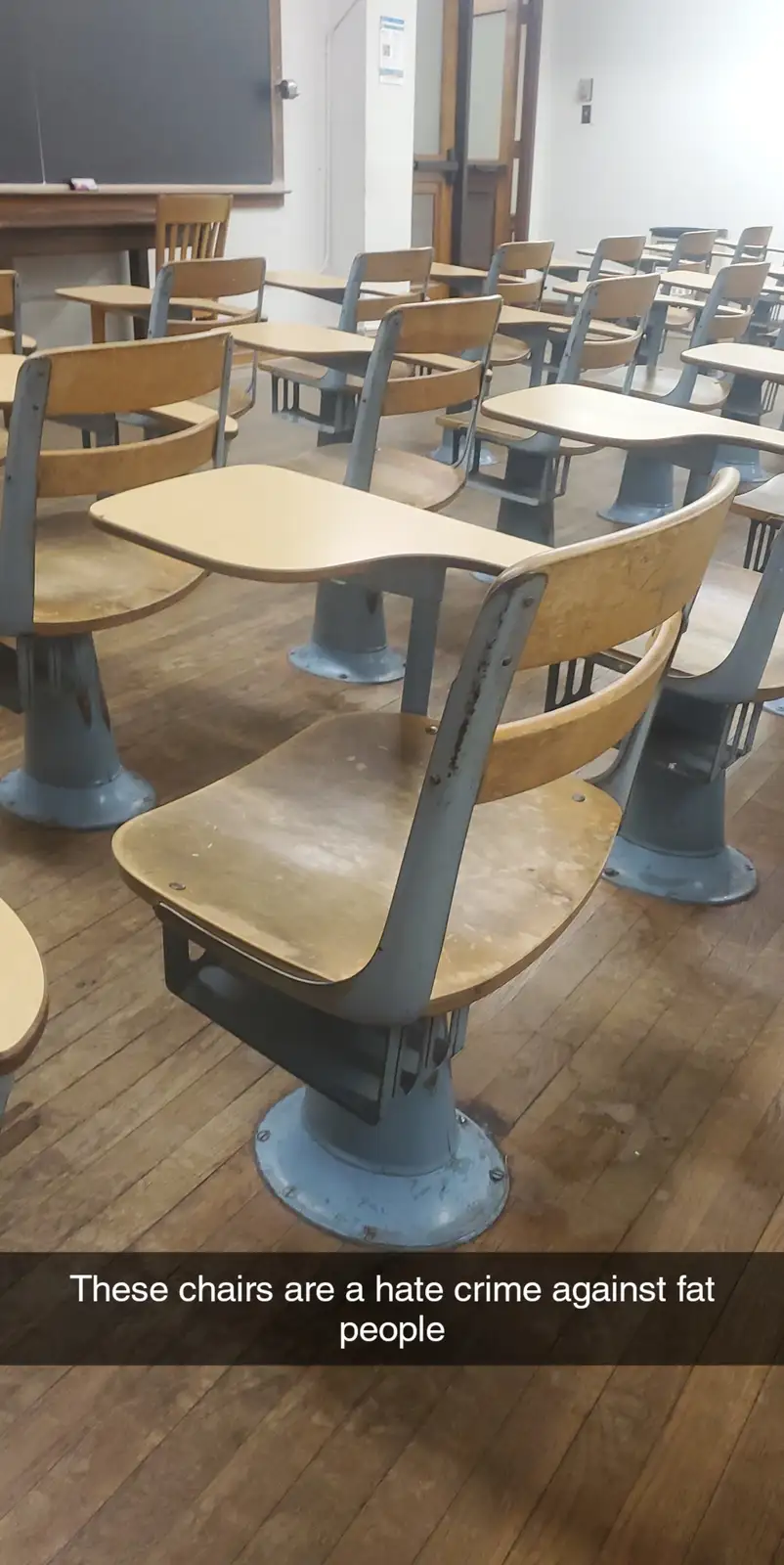 An empty classroom with rows of vintage wooden and metal school desks and chairs on a wooden floor, with a blackboard…