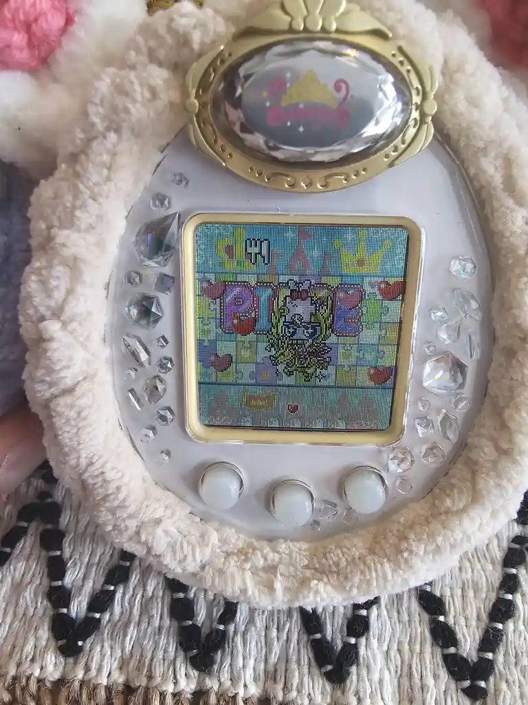 A heavily customized Tamagotchi with a fluffy white trim and numerous glittery rhinestones, displaying the word 'PIGE' in…