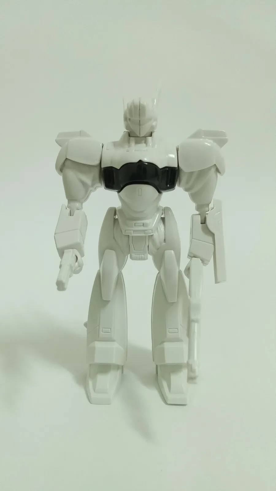 A white plastic robot action figure stands against a plain white background, holding a small gun in its right hand and a…