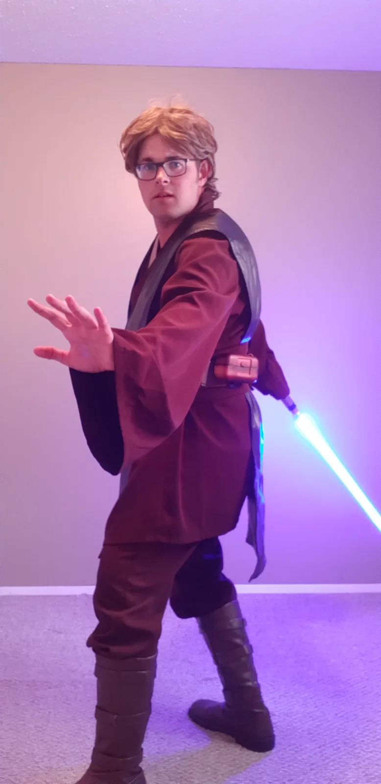 A person in a brown Jedi robe and glasses strikes a lightsaber stance indoors, holding a glowing blue lightsaber in one hand…