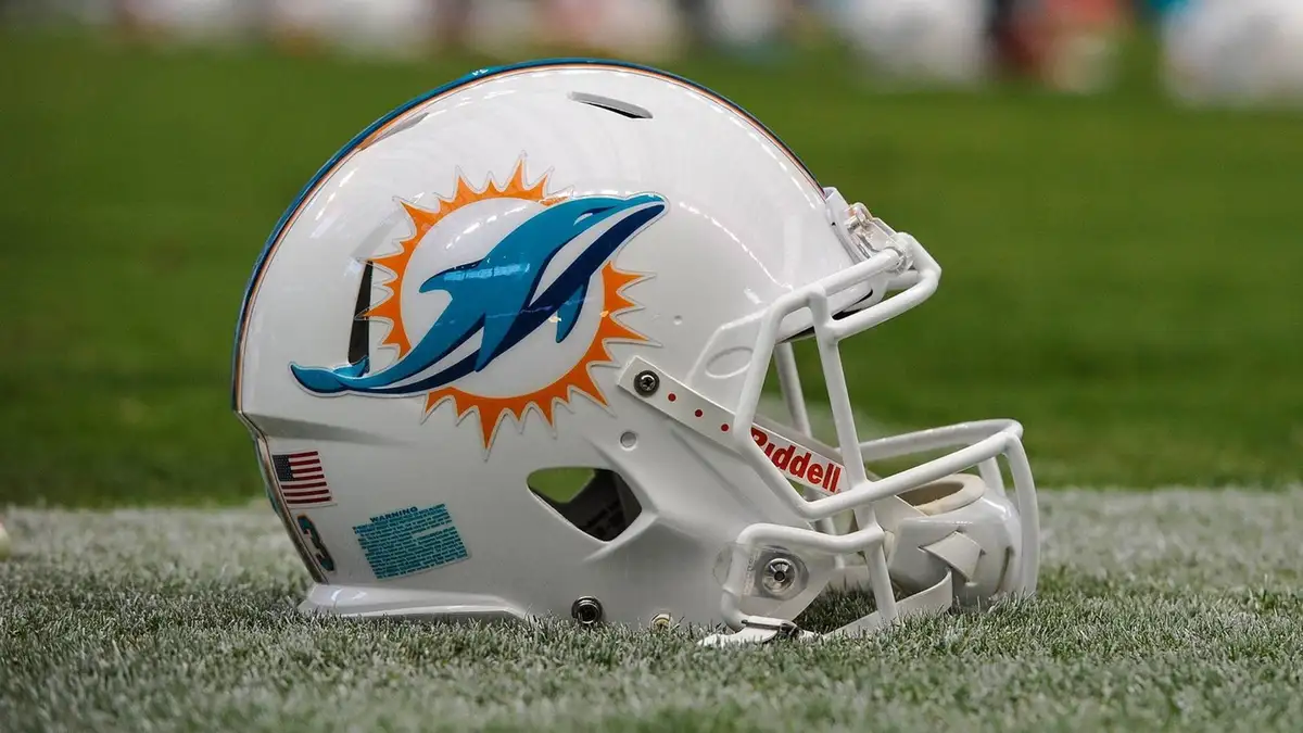 A white Miami Dolphins NFL football helmet with a blue dolphin logo and orange sunburst design rests on green turf. The…