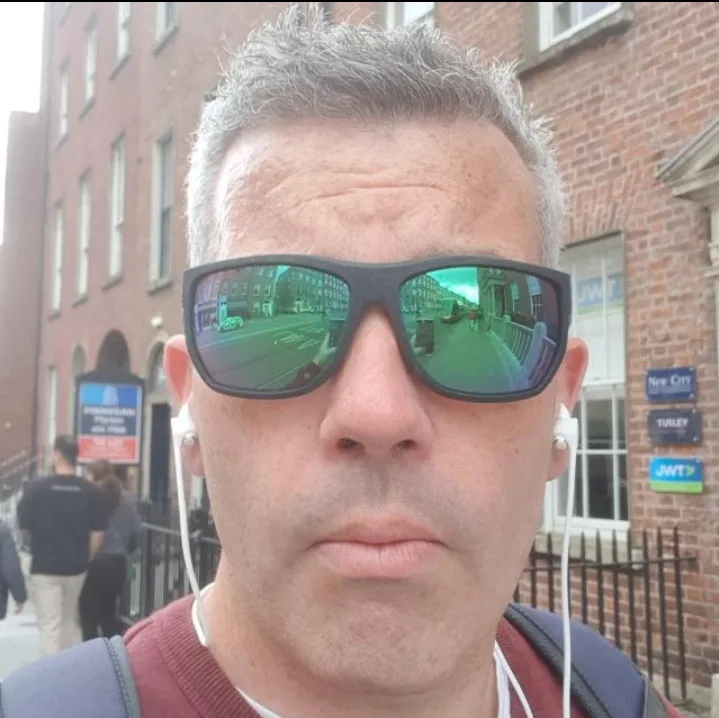 A man with short gray hair wears large black sunglasses and white earbuds while taking a selfie. The reflection in his…