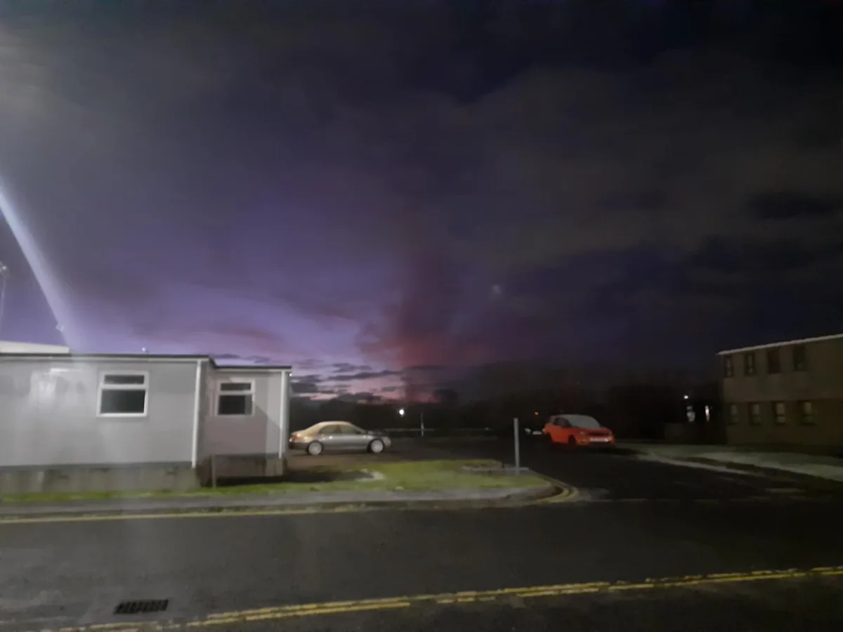 A nighttime photo of a residential area with two parked cars and buildings under a dark sky with purple and red clouds. The…