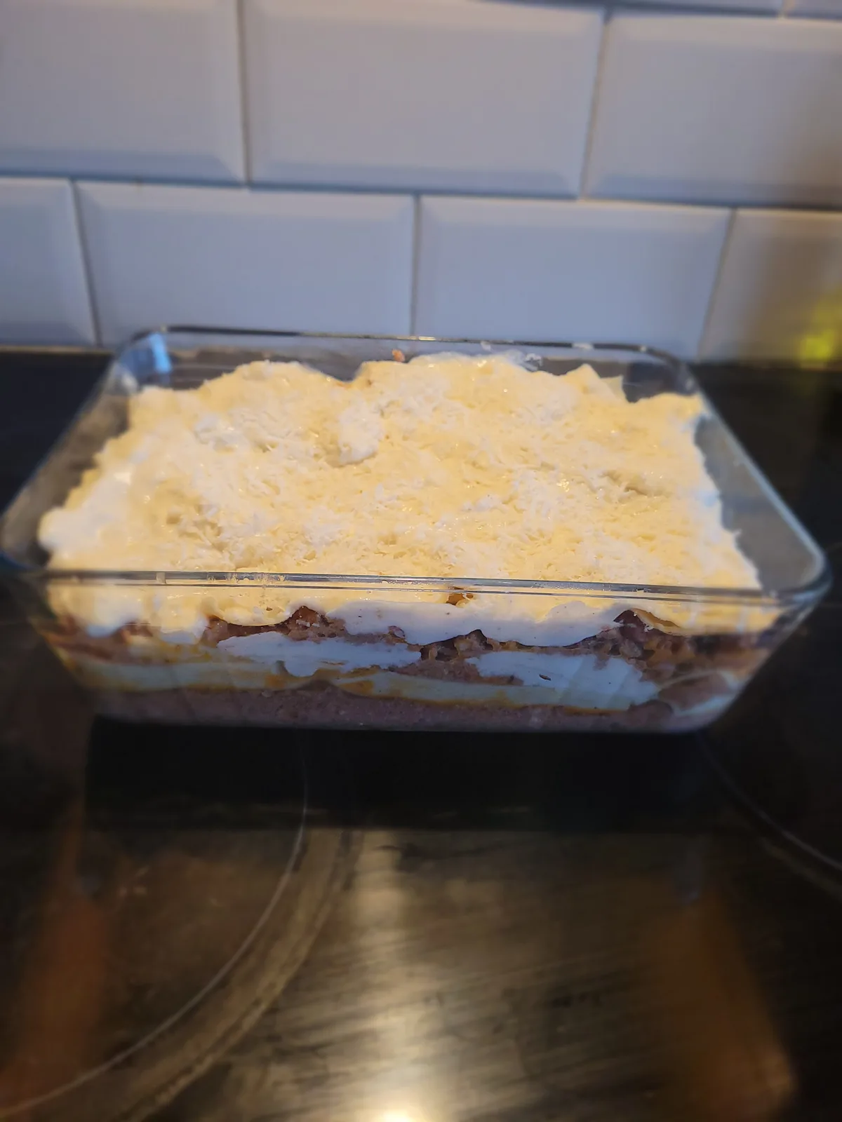 A clear glass baking dish sits on a dark stovetop, containing a layered lasagna with visible pasta, sauce, and white cream…