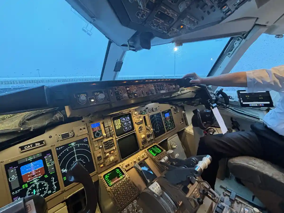 A photo from inside an airplane cockpit during rainy weather, showing a pilot's hand on the control yoke. The cockpit is…