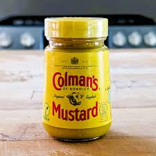 A yellow glass jar of Colman's Mustard sits on a wooden countertop with a stove in the background. The label clearly reads…