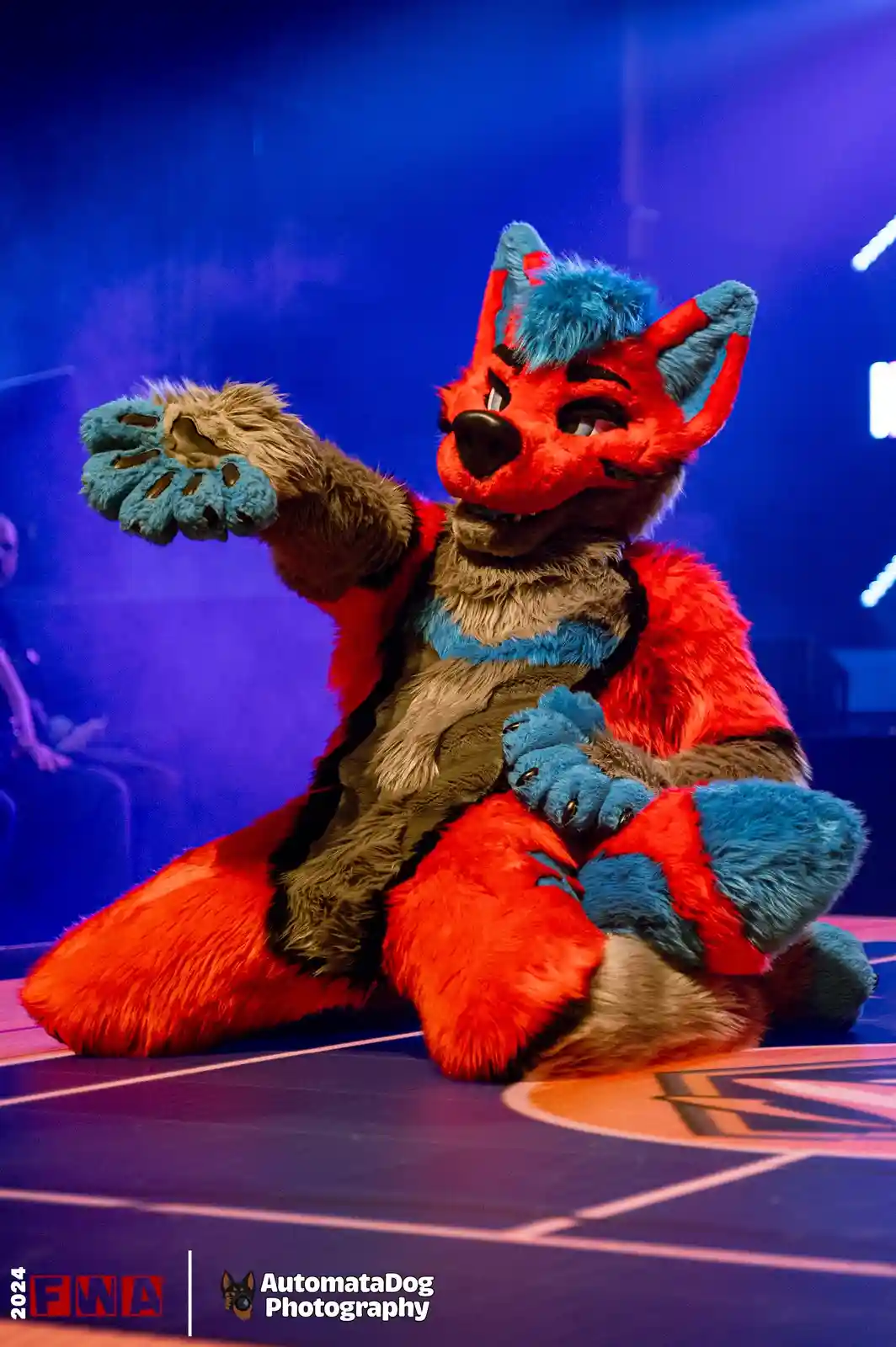 A person in a detailed red, blue, and brown fox fursuit sits on a stage with a smug, confident expression, one paw raised…