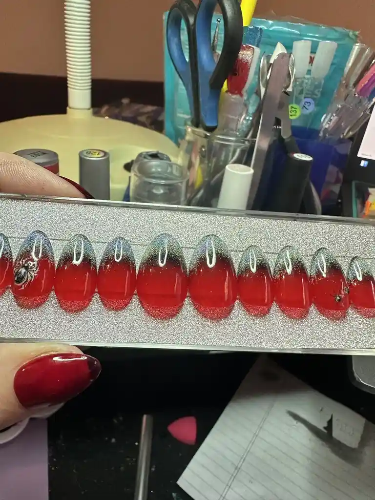 A hand holds a tray of artificial fingernails with a red-to-silver ombre glitter design and white crescent moons. Two nails…