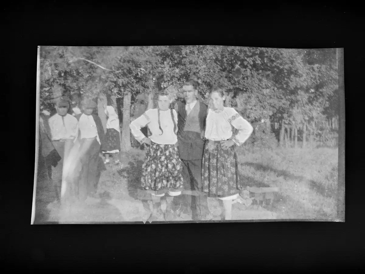 A grainy, black-and-white vintage photograph shows a group of people outdoors, with three individuals in the foreground…