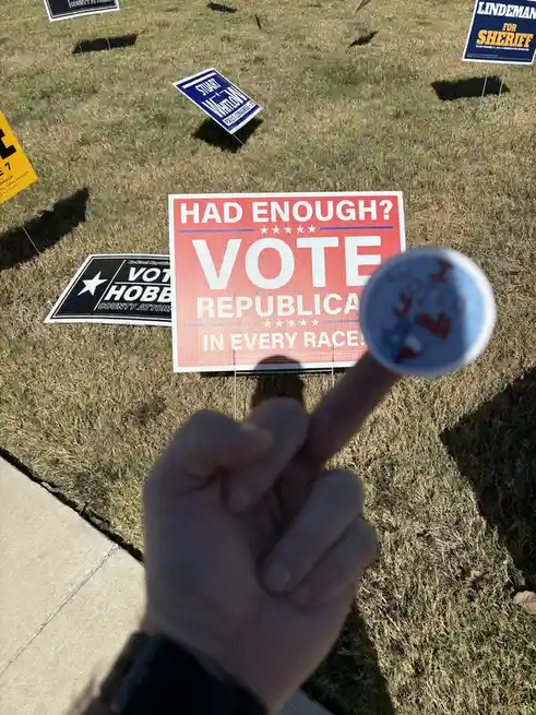 A person's finger, adorned with a small blue sticker that reads 'I voted y'all', points toward a red political yard sign…