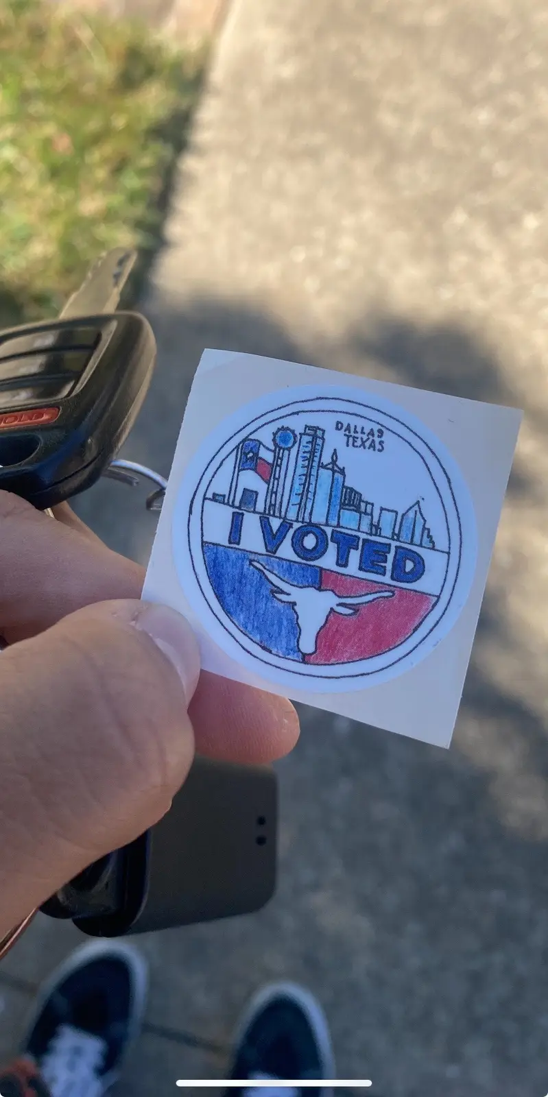 A hand holds a hand-drawn 'I VOTED' sticker featuring the Dallas, Texas skyline and a longhorn silhouette in red and blue…