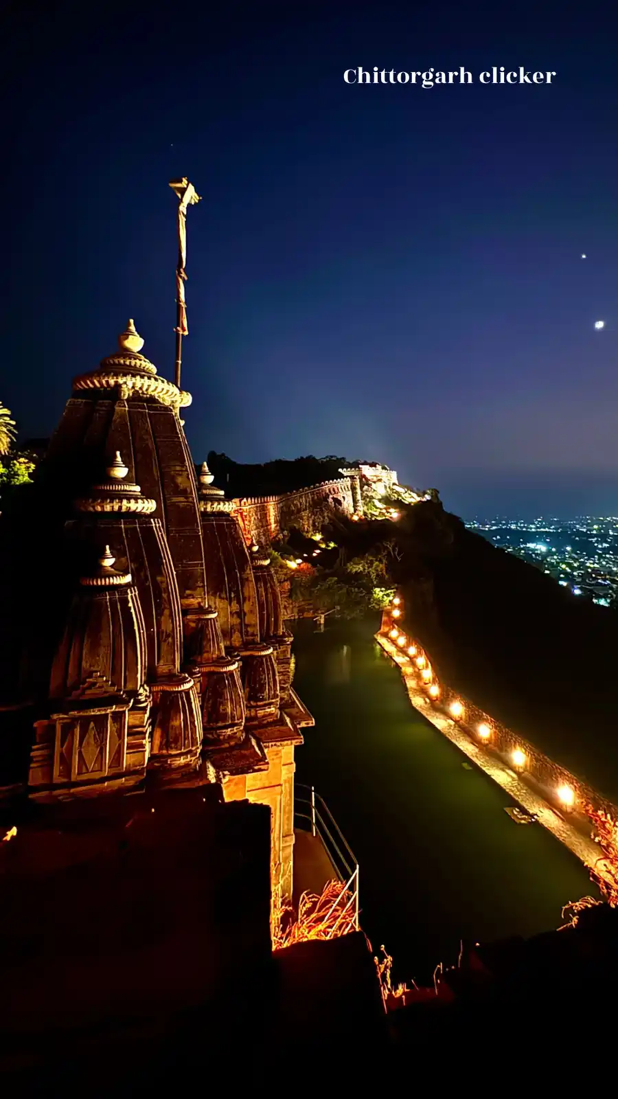 A nighttime photograph of the Chittorgarh Fort in India, showcasing illuminated stone architecture, a lit pathway along a…