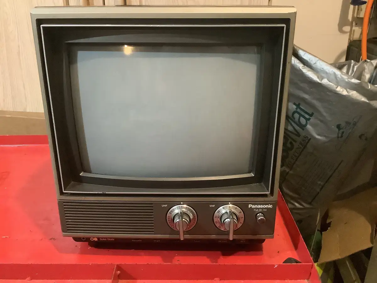 A vintage Panasonic CRT television with two dials sits on a red surface, its screen blank and gray. The overlaid text reads…