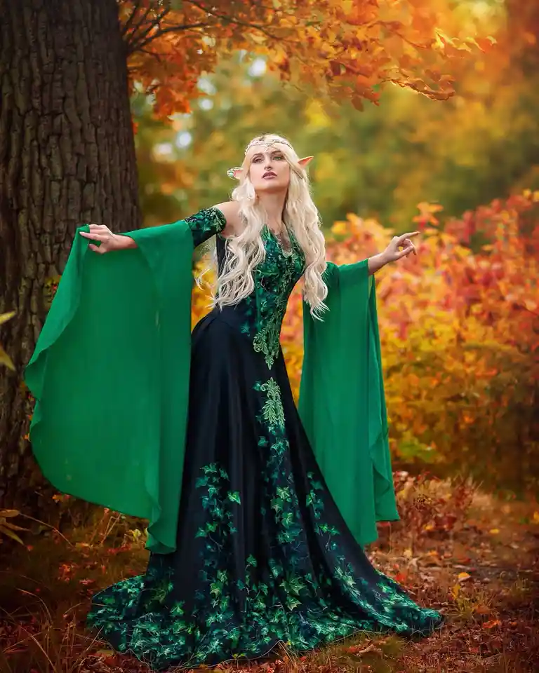 A woman with long blonde hair, pointed elf ears, and a gold headband stands in an autumn forest wearing a long black dress…