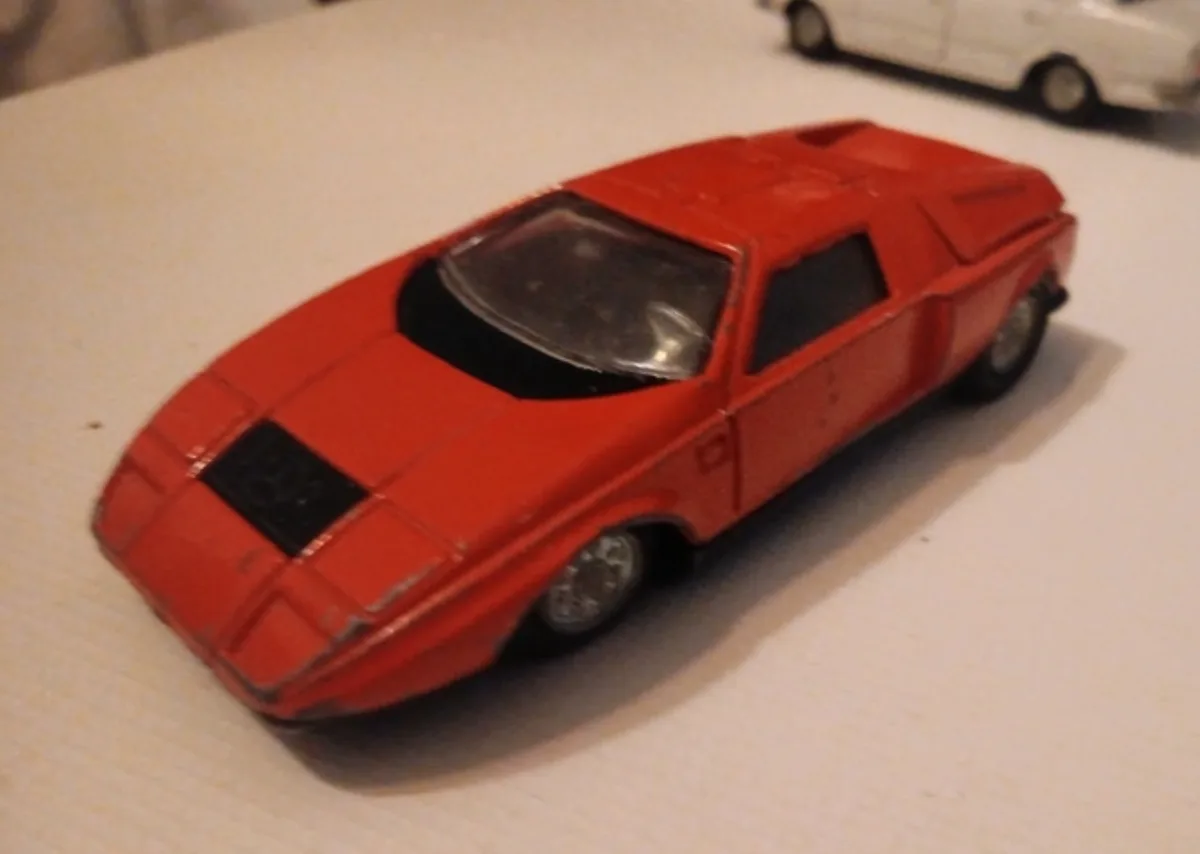 A red toy car with a futuristic, angular design sits on a white surface, with another toy car blurred in the background. The…