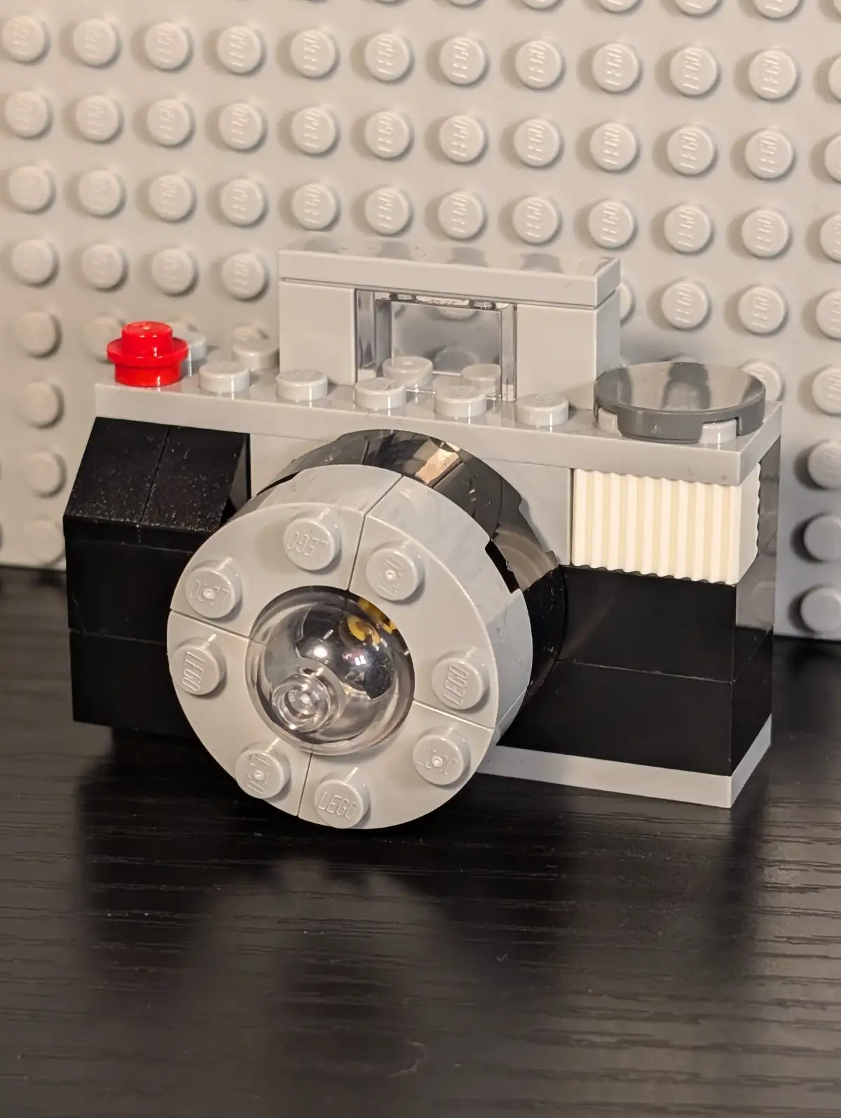 A LEGO-built camera model, constructed from gray, black, and white LEGO bricks, sits on a dark wooden surface against a gray…