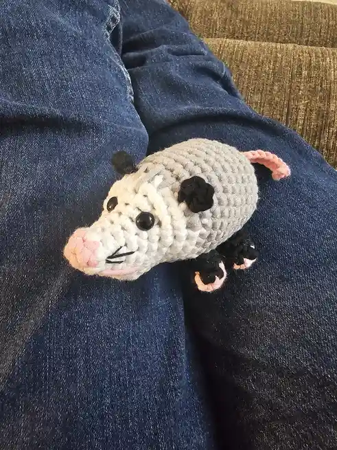 A small, crocheted opossum plush toy with gray, white, black, and pink yarn rests on a person's blue denim-clad lap. The toy…