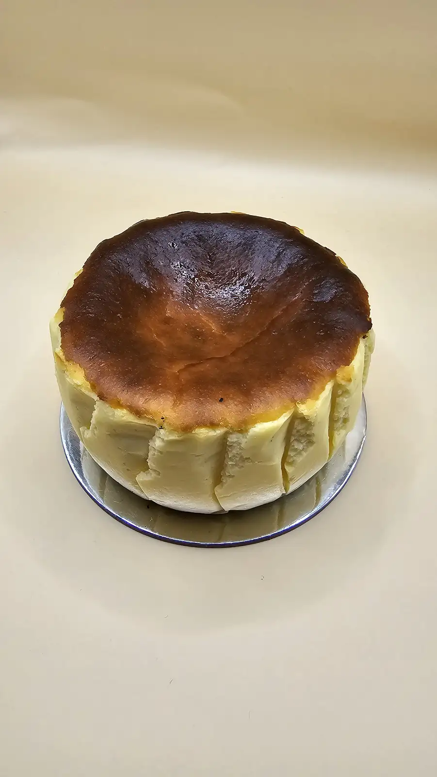 A round baked cheesecake with a golden-brown, slightly cracked top sits on a silver plate against a plain off-white…