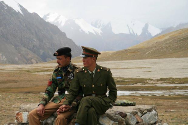 Two soldiers in military uniforms sit side by side on a rocky outcrop in a vast, barren mountainous landscape with…