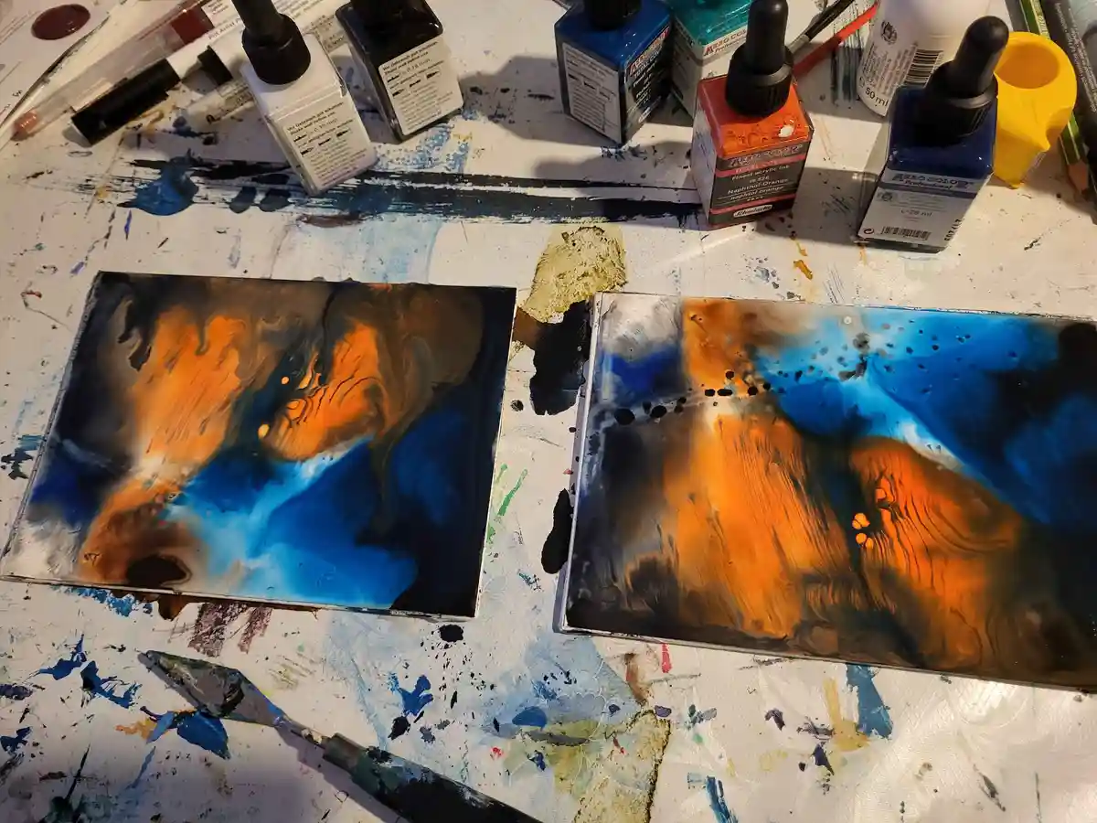 Two abstract acrylic paintings on rectangular panels sit side-by-side on a paint-splattered white artist's table, surrounded…