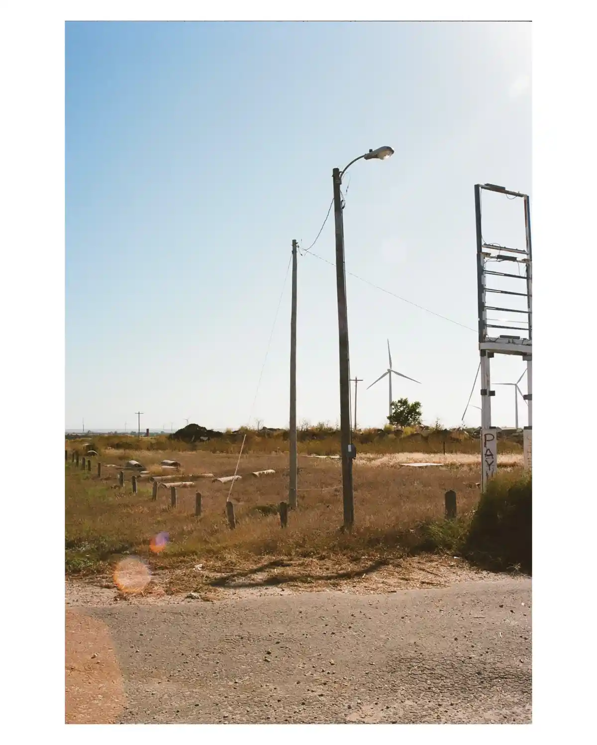 A desolate roadside scene with dry grass, wooden posts, and a broken streetlight pole under a clear blue sky, with wind…