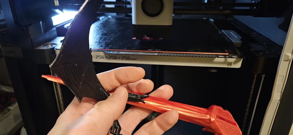 A hand holds a 3D-printed bat wing with a red shaft and black wing, in front of a 3D printer bed labeled '256 x 256 x…