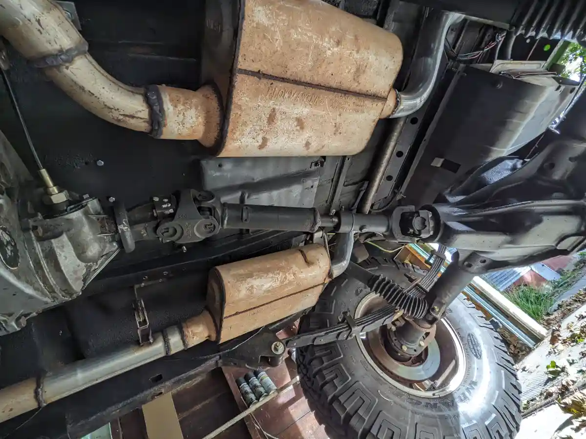 A close-up view of the underside of a lifted off-road vehicle, showing two rusty Flowmaster exhaust mufflers, a driveshaft…