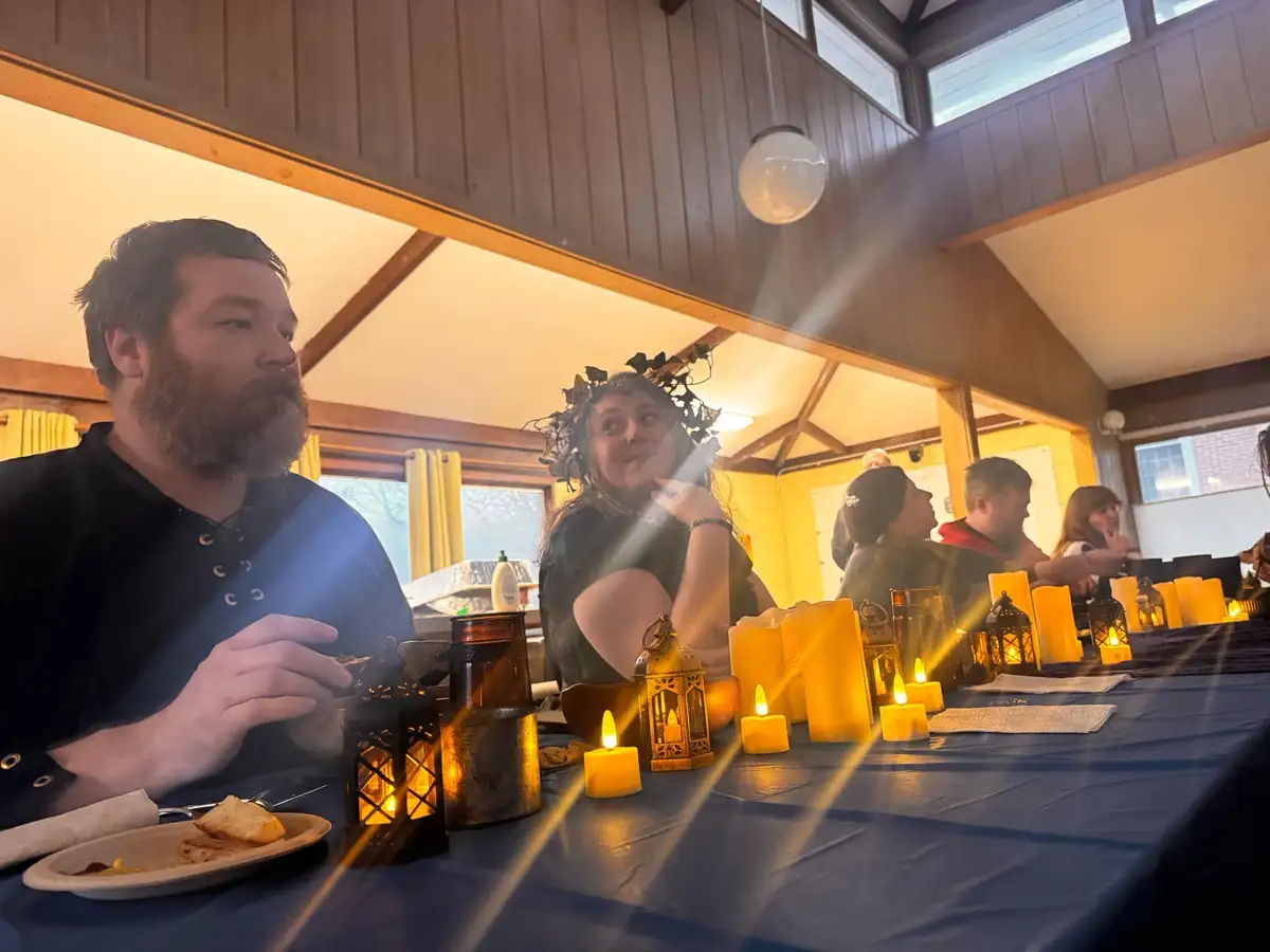 A group of people are seated at a long table covered with a blue tablecloth, surrounded by numerous lit candles and…