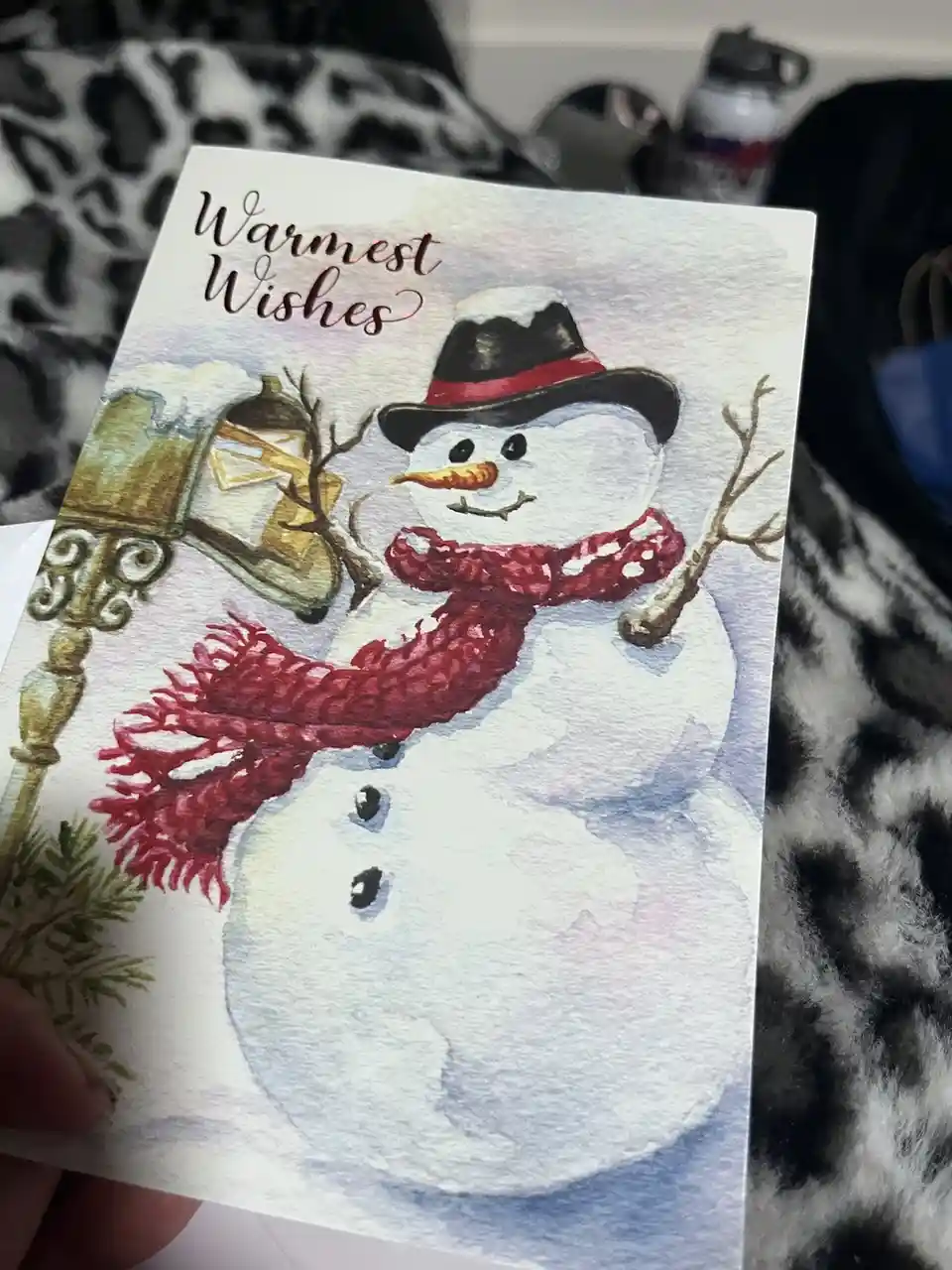 A hand holds a watercolor holiday card featuring a cheerful snowman wearing a black top hat with a red band, a red knitted…