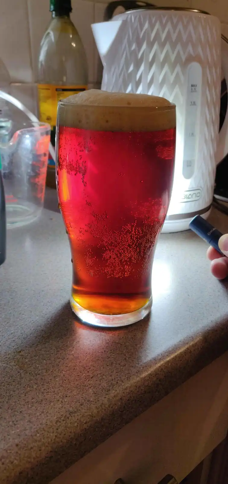 A tall glass filled with a red, carbonated liquid and a foamy head sits on a kitchen counter, with a hand holding a vape pen…