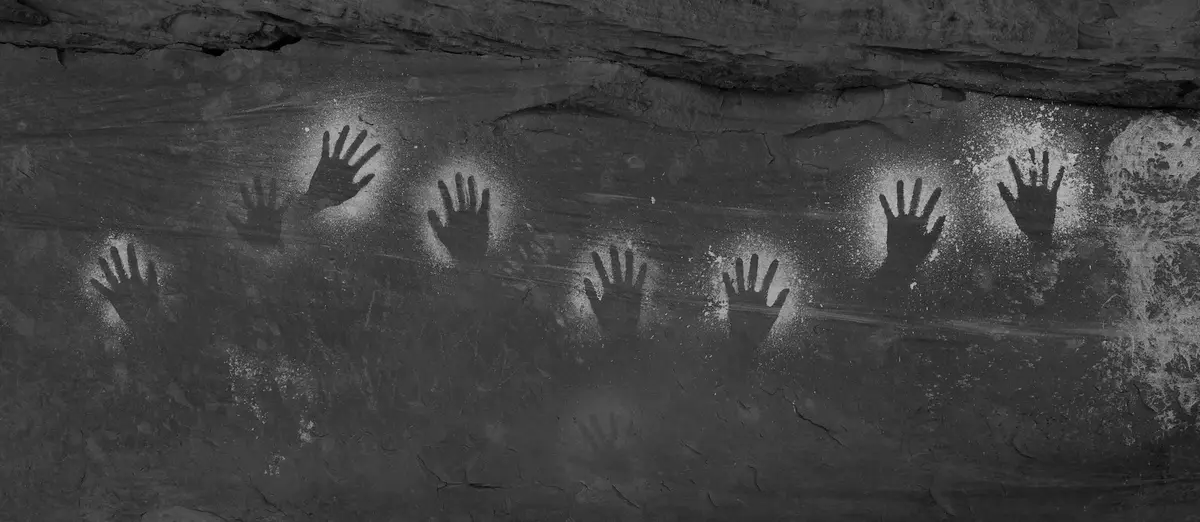 A black-and-white image shows multiple handprints pressed into a dark, textured rock surface, resembling ancient…