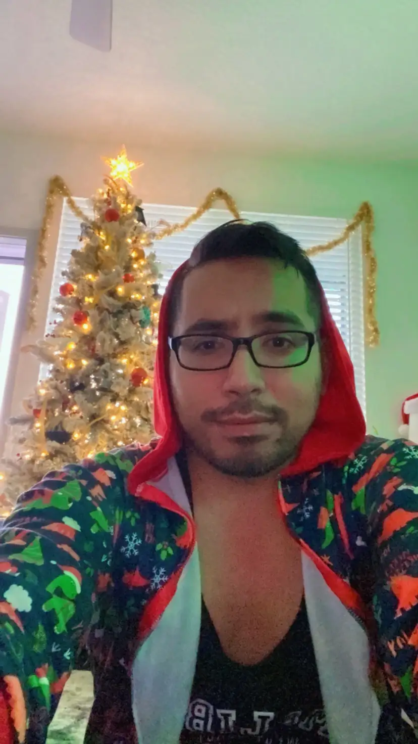 A man wearing glasses and a colorful holiday-themed hoodie with the hood up stands in front of a lit Christmas tree…