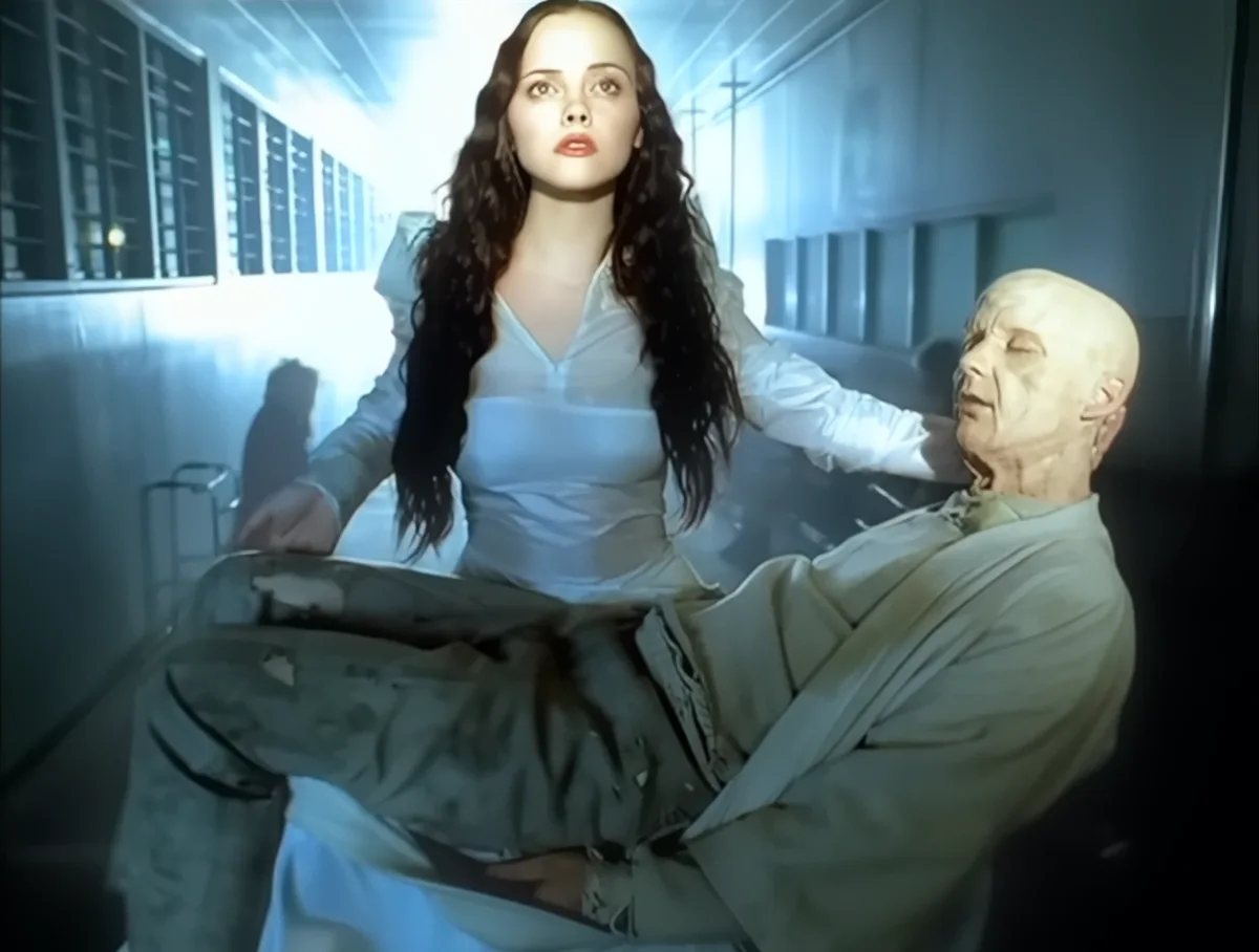 A woman with long dark hair in a white shirt sits beside a bald man in a light-colored suit who is slumped over, appearing…