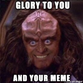 A close-up of a bald, pink-skinned alien with long dark hair, wide blue eyes, and a toothy grin. The image has white, bold…