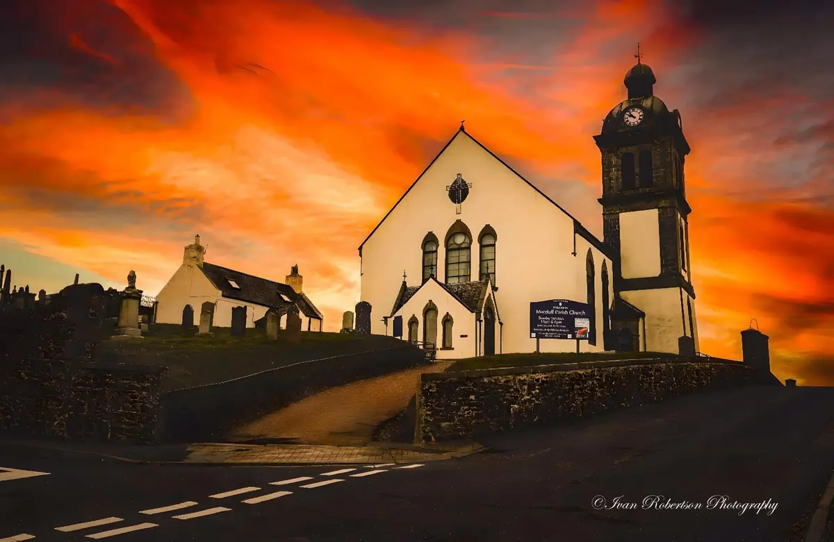 A picturesque church with a clock tower stands under a dramatic, fiery orange and red sunset sky. A stone wall and a road…