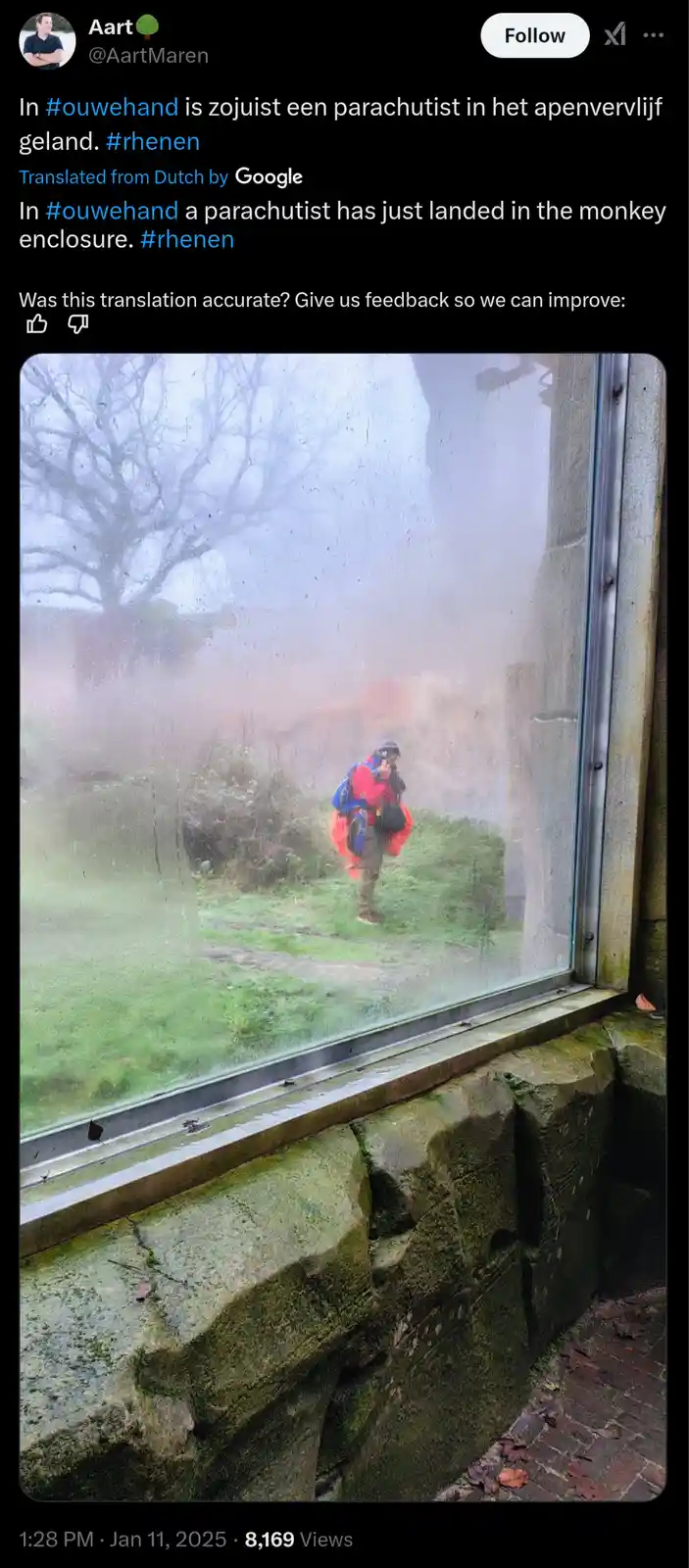 A person in a red and blue parachute suit is seen walking outside through a rain-splattered glass window, viewed from inside…